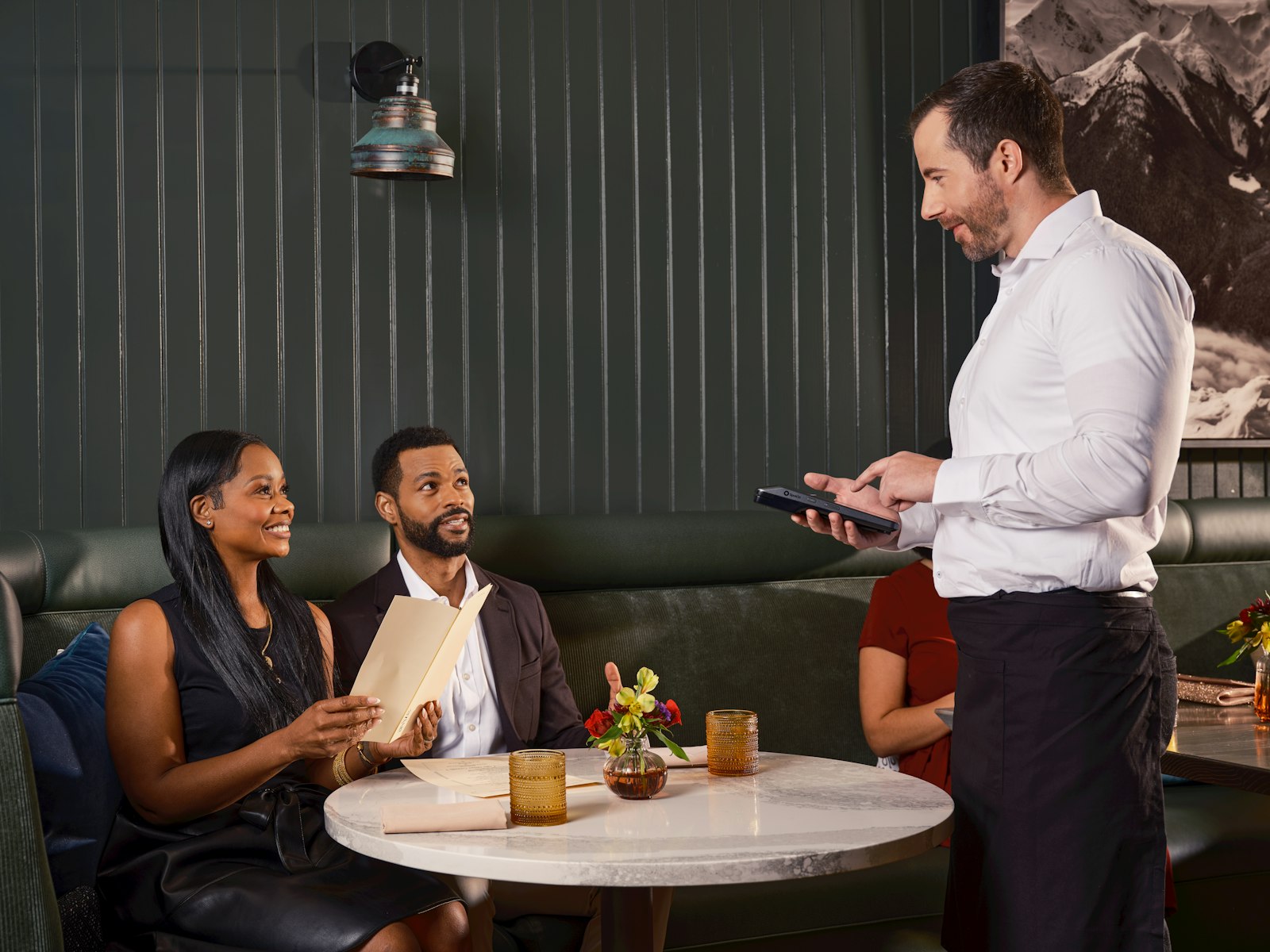 Un serveur prend la commande d'un couple installé en banquette, sa tablette à la main.